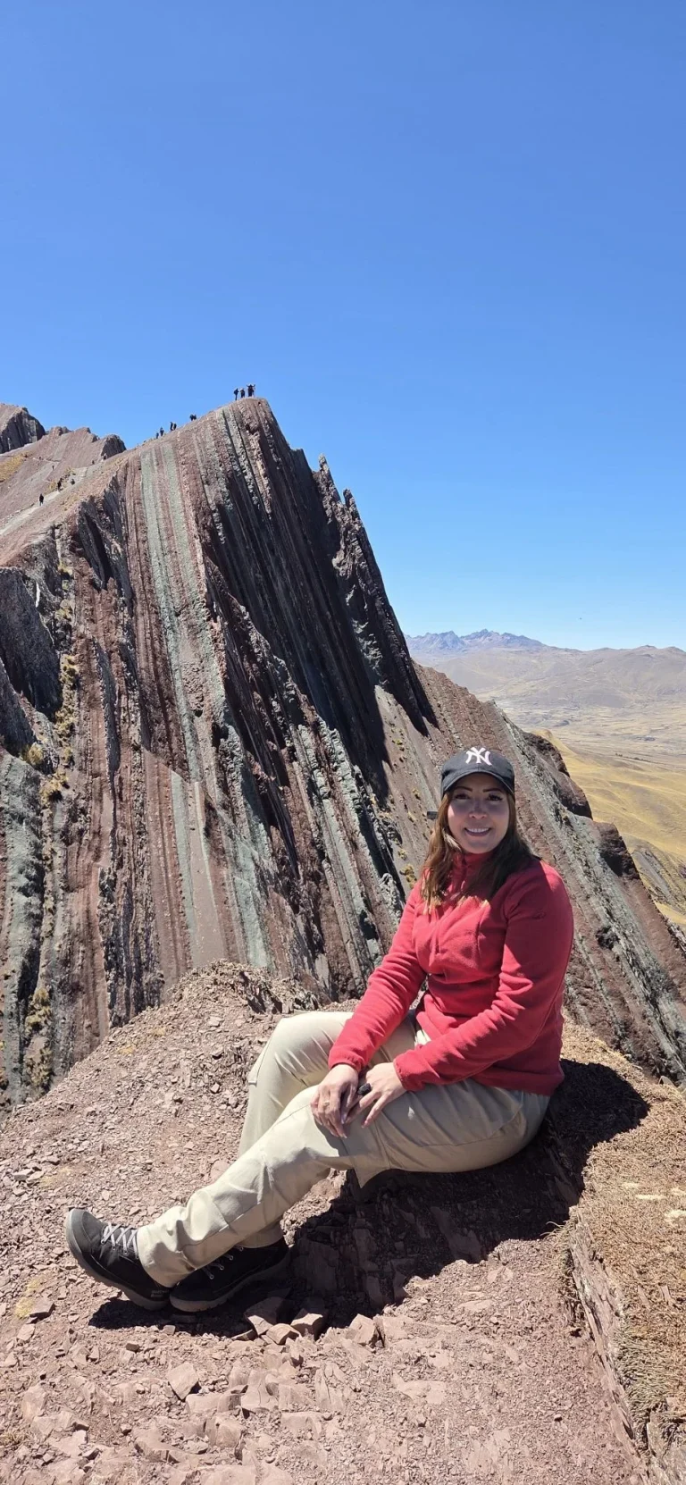 Hiker sitting on the ridge of Pallay Punchu with the striped main peak and hikers on the summit behind